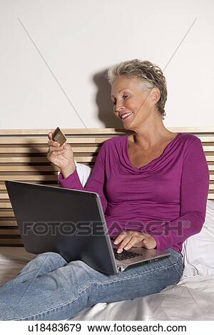 Stock Photo - Older woman shopping online on bed. Fotosearch