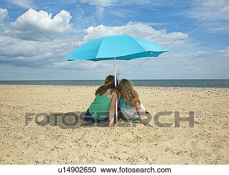 Stock Image - Sisters sitting under umbrella on beach. Fotosearch