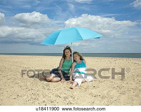 Stock Image - Sisters sitting under umbrella on beach. Fotosearch