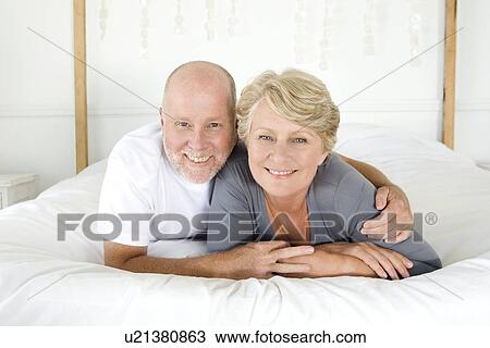 Stock Image - Smiling older couple laying on bed. Fotosearch