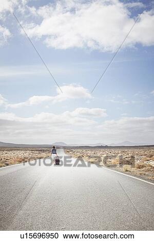 Stock Image - Woman rolling luggage on rural road. Fotosearch