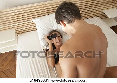 Stock Image - Woman taking video of boyfriend in bed. Fotosearch
