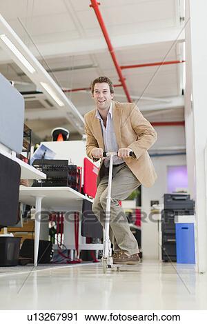Stock Image - Businessman riding scooter in office. Fotosearch