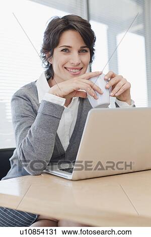 Stock Image - Businesswoman drinking coffee at desk. Fotosearch