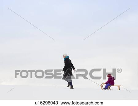 Stock Image - Mother pulling daughter on sled in snow. Fotosearch
