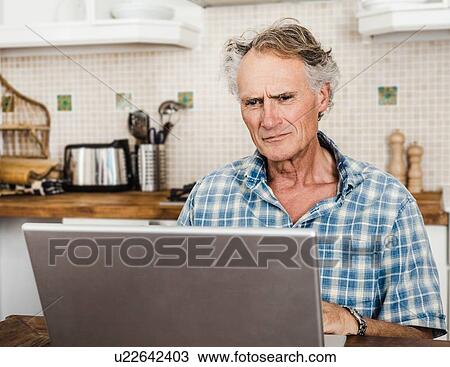 Stock Image - Older man using laptop in kitchen. Fotosearch