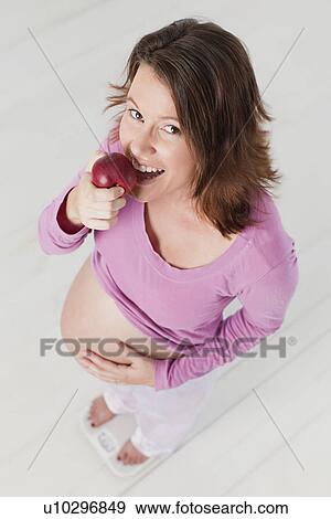Stock Photo - Pregnant woman eating apple on scale. Fotosearch