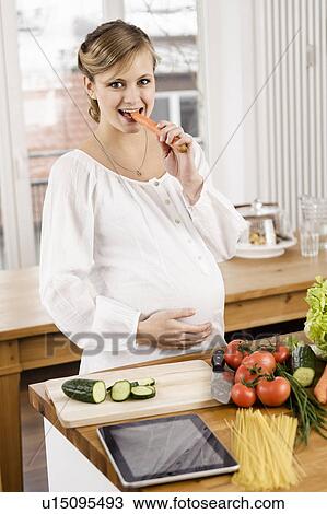 Stock Image - Pregnant woman eating carrot in kitchen. Fotosearch