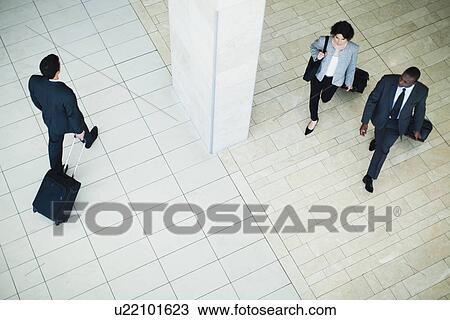Stock Image - Business people rolling luggage in lobby. Fotosearch