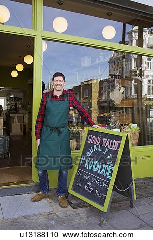 Stock Image - Smiling grocer standing outside store. Fotosearch