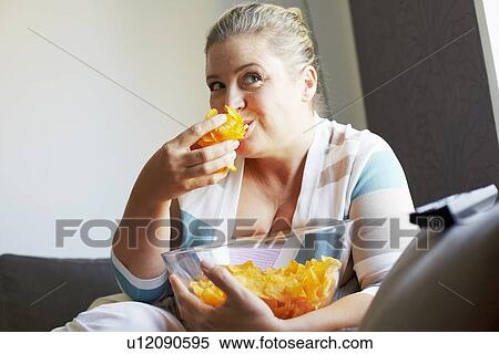 Stock Photography - Smiling woman eating chips on sofa. Fotosearch