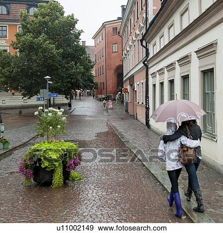 Stock Photo - Two female friends holding an umbrella in rain on a street, Uppsala, Sweden. Fotosearch