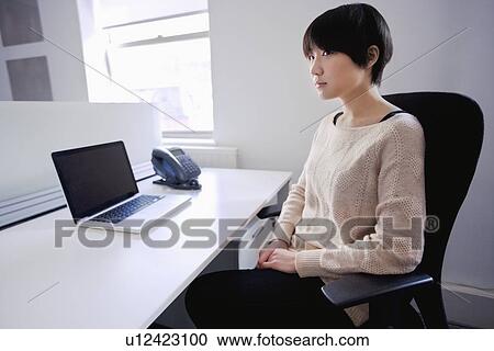 Stock Image - Asian woman sitting at desk with laptop while looking away. Fotosearch