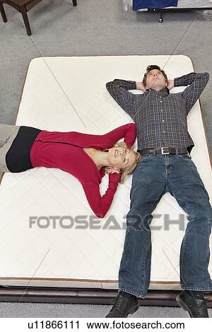 Stock Image - Friends lying on mattress with hands behind head in furniture store. Fotosearch