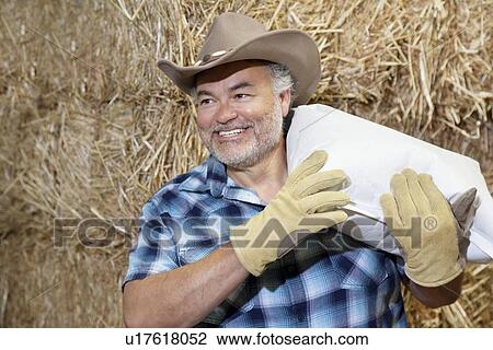 Stock Image - Happy mature cowboy carrying sack on shoulder while looking away. Fotosearch