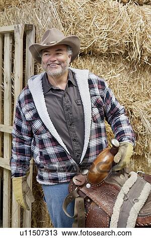 Stock Image - Happy mature cowboy holding saddle. Fotosearch