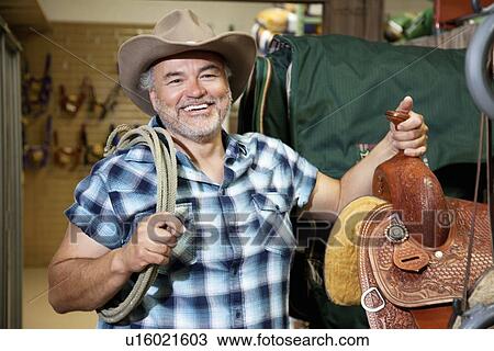 Stock Image - Happy mature cowboy with saddle and rope in feed store. Fotosearch