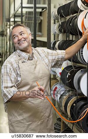 Stock Photo - Happy mature salesperson standing by electrical wire spool while looking away in hardware store. Fotosearch
