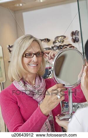 Happy senior woman trying on glasses while optician holding hand mirror View Large Photo Image Stock Image - Happy senior woman trying on glasses while optician holding hand mirror. Fotosearch