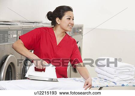 Stock Image - Happy young female employee ironing while looking away in Laundromat. Fotosearch