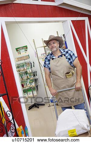 Stock Image - Mature cowboy pushing cart with sack in feed store. Fotosearch