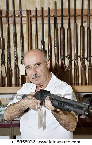 Stock Image - Mature gun merchant with rifle looking away. Fotosearch