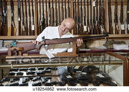 Stock Image - Mature gun shop owner looking at rifle in store. Fotosearch