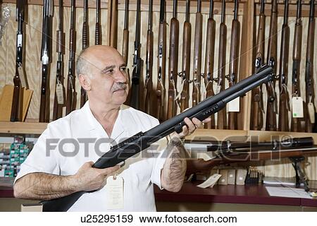 Mature gun store owner looking at weapon in shop View Large Photo Image Stock Photo - Mature gun store owner looking at weapon in shop. Fotosearch