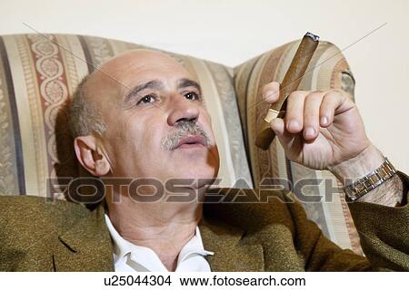 Mature man smoking cigar while relaxing on armchair View Large Photo Image Picture - Mature man smoking cigar while relaxing on armchair. Fotosearch
