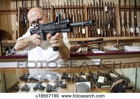 Stock Image - Mature merchant aiming with rifle in gun shop. Fotosearch