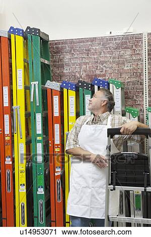 Stock Image - Mature salesperson standing by multicolored ladders in hardware store. Fotosearch