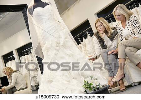 Stock Image - Mother and daughter sitting on sofa while looking at elegant wedding dress in bridal store. Fotosearch