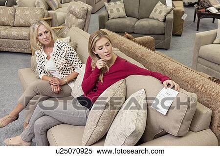 Stock Photography - Mother and daughter sitting on sofa while looking at price tag in furniture store. Fotosearch