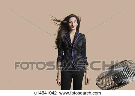 Stock Image - Portrait of a beautiful woman standing while fan blowing her hair over colored background. Fotosearch