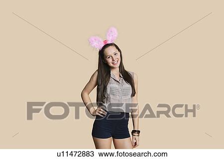 Portrait of a beautiful young woman wearing bunny ears with hands on hips over colored background View Large Photo Image Stock Image - Portrait of a beautiful young woman wearing bunny ears with hands on hips over colored background. Fotosearch