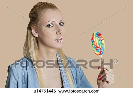 Stock Photography - Portrait of a blond woman with sprinkled lips holding lollipop over colored background. Fotosearch