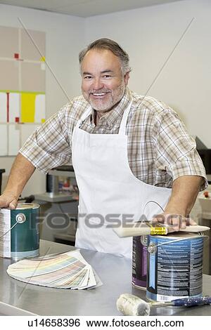 Stock Photograph - Portrait of a cheerful mature store clerk with paint cans and brush in hardware shop. Fotosearch