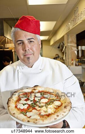 Picture - Portrait of a confident chef wearing headgear while holding pizza in commercial kitchen. Fotosearch
