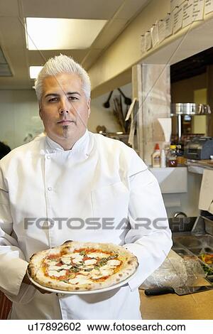 Stock Image - Portrait of a confident mid adult chef holding pizza in commercial kitchen. Fotosearch