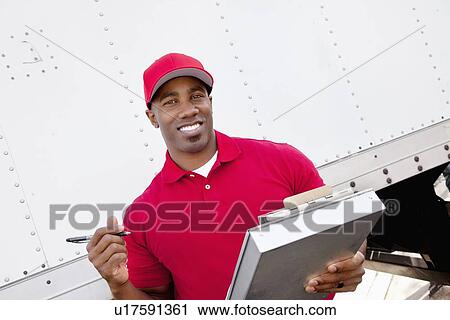 Stock Image - Portrait of a happy African American man holding clipboard with delivery truck in background. Fotosearch