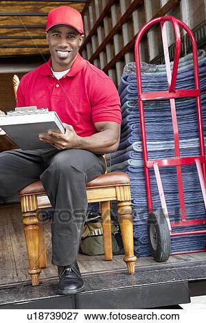 Stock Photo - Portrait of a happy delivery man sitting on chair with clipboard. Fotosearch