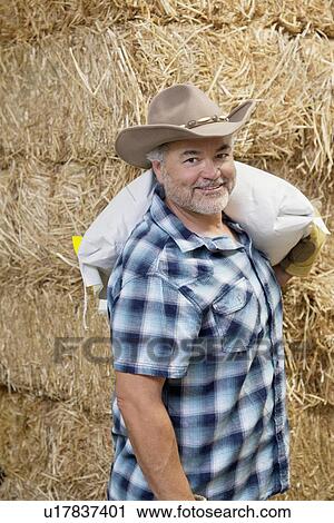 Stock Image - Portrait of a happy mature cowboy carrying sack on shoulder. Fotosearch