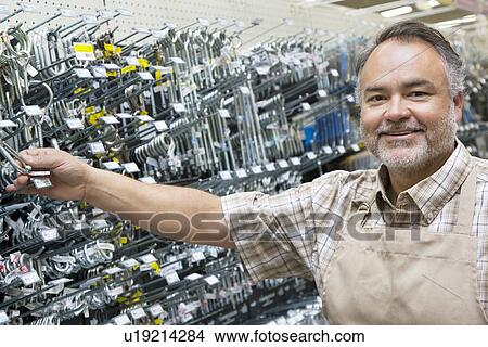 Picture - Portrait of a happy mature salesperson holding metallic equipment in hardware store. Fotosearch