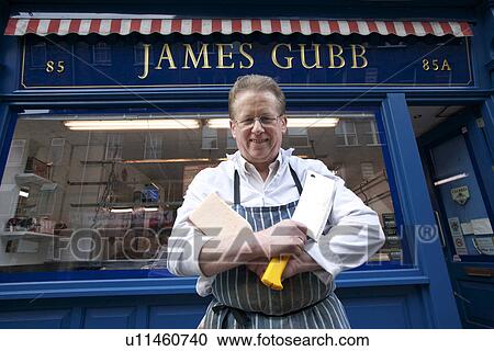Stock Image - Portrait of a happy senior butcher standing with crossed cleavers in hands outside shop. Fotosearch