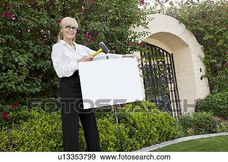 Portrait of a happy senior female agent hammering sign board in lawn View Large Photo Image Stock Photo - Portrait of a happy senior female agent hammering sign board in lawn. Fotosearch
