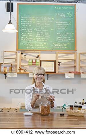 Stock Image - Portrait of a happy senior merchant with spice jar at counter in store. Fotosearch