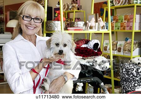 Stock Image - Portrait of a happy senior woman carrying dog in pet shop. Fotosearch