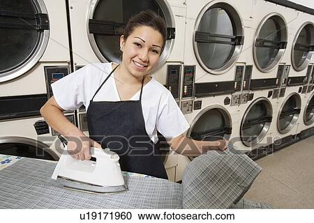 Stock Image - Portrait of a happy woman wearing apron ironing in front of washing machines. Fotosearch