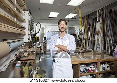 Stock Photo - Portrait of a happy young craftsman standing with arms crossed in workshop. Fotosearch