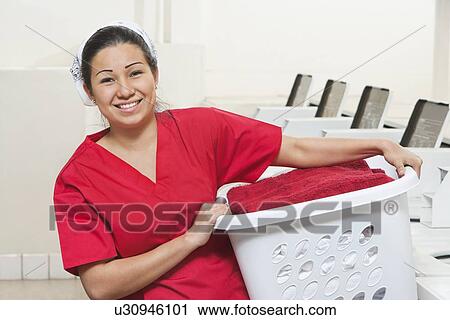 Stock Image - Portrait of a happy young female employee carrying laundry basket. Fotosearch